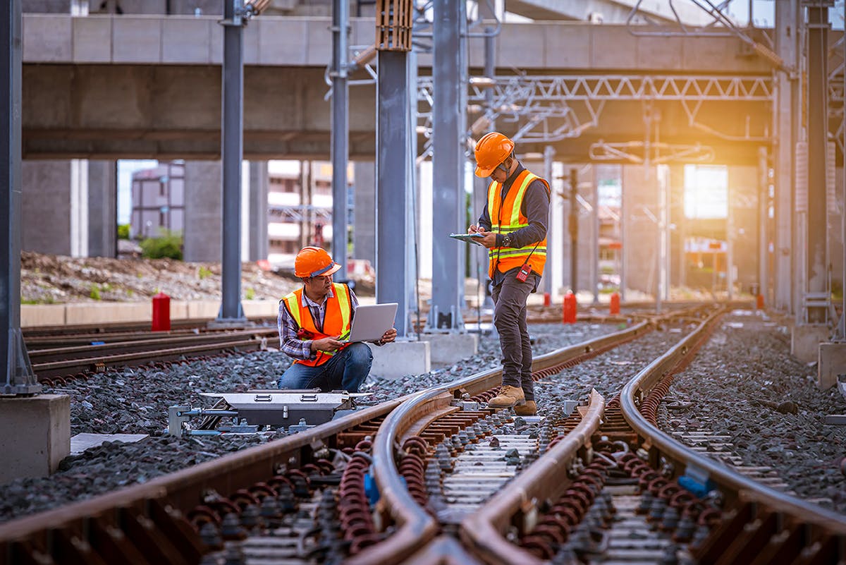 Two engineers in safety uniforms inspect a railway using a laptop for enterprise asset management, linear assets, big data system, enterprise content management, digital asset management, and records management.